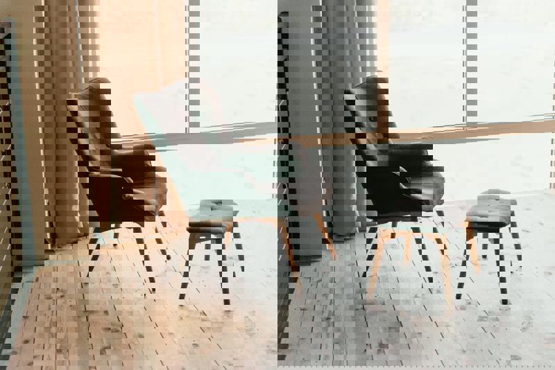 En elegant mörkgrå fåtölj med träben och matchande fotpall står på ett ljust trägolv, placerad vid ett stort fönster med utsikt och ljusa gardiner som ramar in rummet.