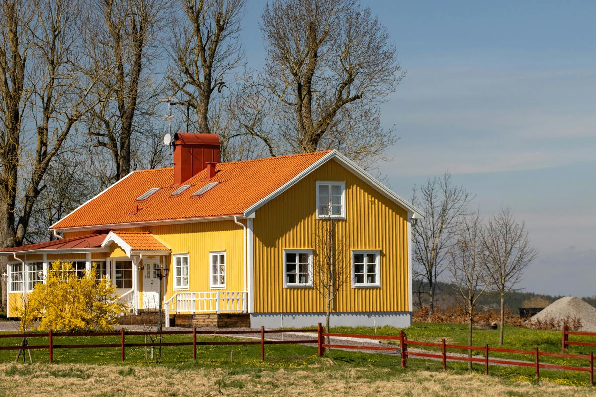 Traditionellt svenskt hus målat i gult med vita knutar och rött tak. Huset står i en lantlig miljö med gräsmatta, träd och blommande buskar i förgrunden, och blå himmel i bakgrunden.