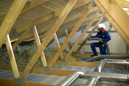 Person i skyddskläder sprutar isoleringsmaterial på en vind med träbjälkar och ventilationsrör.