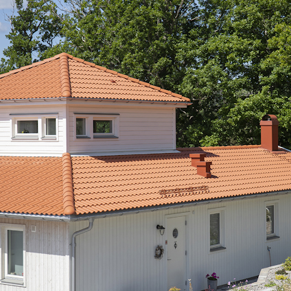 Vitt hus med orange tegeltak, vit panel och tre fönster. Grönska i bakgrunden under en klar himmel.