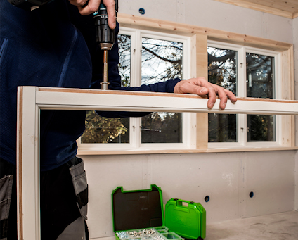 En person monterar en karmhylsa i en fönsterkarm som står löst. I bakgrunden syns fönster och två förvaringslådor på golvet.