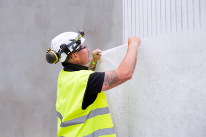 Person i skyddsväst och hjälm applicerar vit väggbeläggning med roller.