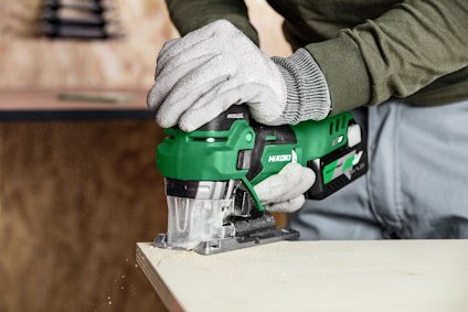En person håller en grön sticksåg och sågar i en träplanka. Sawdust faller från kanten.